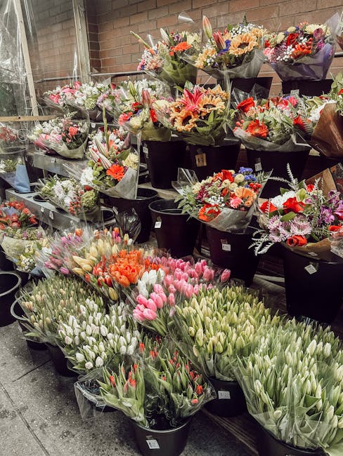 A variety of fresh flower bouquets and arrangements displayed outdoors on tiered black shelves at a florist shop, featuring colorful blooms such as pink, orange, red, purple, and white tulips, daffodi
