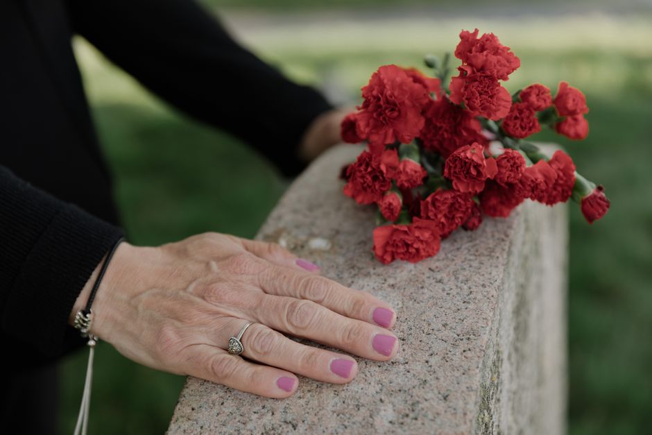 A pair of hands resting on a weathered stone monument outdoors, with a red floral arrangement of carnations placed on top. The person has neatly manicured nails painted pink, wears a silver ring with 