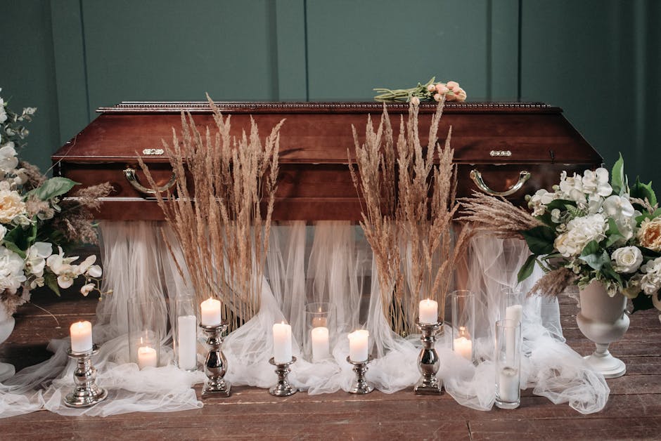 A wooden casket is decorated with floral arrangements featuring white roses, orchids, and soft pink blossoms on either side, set against a green wall. Behind the casket, there are tall, dried pampas g