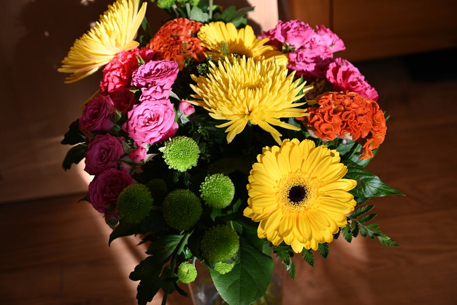 A vibrant floral bouquet featuring yellow gerbera daisies, pink roses, orange carnations, green chrysanthemums, and yellow chrysanthemums arranged in a clear glass vase. The flowers are freshly cut, w