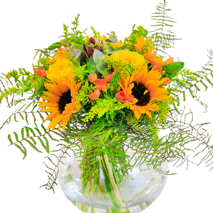With a flourish of colour and warmth, this floral arrangement embodies the joyous spirit of New Addington. Dominant sunflowers gaze outward, their large petals unfurling like rays of sunshine over Fairchildes Avenue, providing a cheerful cascade of yellow around rich, espresso-brown centres. These are gracefully intertwined with buttery yellow garden roses, each flower's petals layered in tight, unfolding spirals akin to the perfection found in the community's cherished gardens. Clusters of red Hypericum berries punctuate the bouquet, sparkling amidst deep green foliage and delicate ferns that seem to spill over the vase's edge. Lacy fillers-likely goldenrod-dot the landscape with miniature yellow blossoms, while slender, blue-green eucalyptus sprigs peek through for a note of cool sophistication. Each element is cradled within a crystal-clear, spherical vase, where brightly coloured stems and fresh water reflect the bouquet's careful craftsmanship by Flowers New Addington florists. Bright daylight exposes every nuance in colour and texture, casting subtle shadows on the table below. This arrangement is ideal for celebrating birthdays, welcoming new beginnings, or simply sharing a little everyday joy-the kind of gift that brings the warmth of New Addington's close-knit community right into your home.